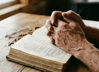 Folded hands resting over an open Bible beside eyeglasses and beads